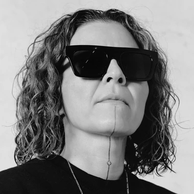 Black-and-white portrait of artist Guadalupe Rosales wearing dark sunglasses, with a vertical line tattoo extending from the lower lip down the neck.