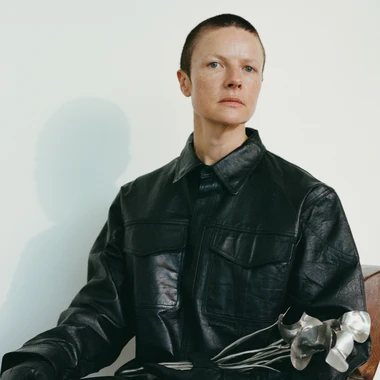 Isabelle Albuquerque portrait with a shaved head and wearing a leather-type long black button up jacket, holding a sword