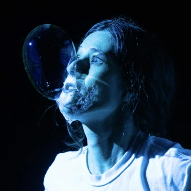 Claire Woolner in blue stage lighting blowing a bubble