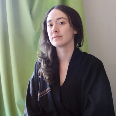 the artist Emi Helfrich wearing a black robe sitting in front of a green curtain