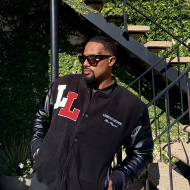 Man in a black Leisure Lounge varsity jacket and sunglasses standing in front of an ivy-covered staircase.