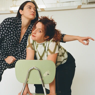 Two members of Finom seated and leaning over a green chair in a bright studio