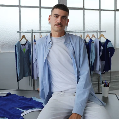 Gabriel Fontana in white jeans and a white shirt with a light blue long-sleeved shirt, posing in front of the uniforms for his work "Multiform"