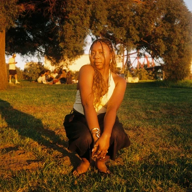 Woman with long braids sitting on grass in a park at golden hour, wearing a white tank top and gold jewelry.