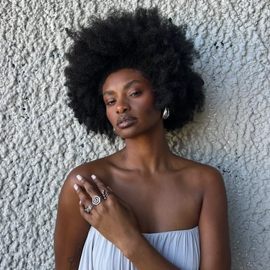 Woman with a full natural afro leaning against a white stucco wall in a lavender strapless dress and silver jewelry.