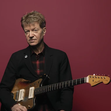 Musician Nels Cline holding a Fender guitar against a deep red background