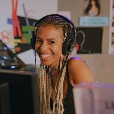 Novena Carmel at a DJ setup wearing headphones, smiling