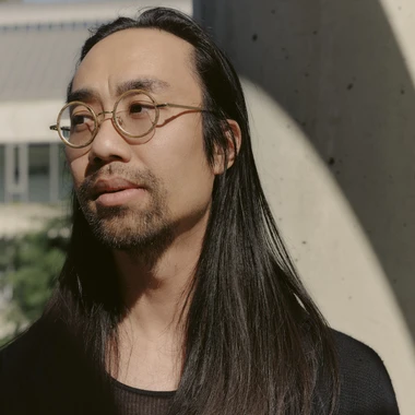Musician Patrick Shiroishi standing outdoors in soft light, long dark hair, wearing glasses