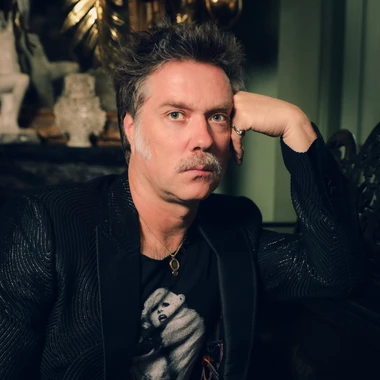 Musician Rufus Wainwright seated in a dimly lit room, resting one hand against their face