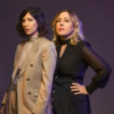 Two members of Sleater-Kinney standing back to back against a purple background