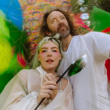 Two members of Sylvan Esso outdoors in colorful clothing, one holding a paintbrush, against a bright painted backdrop