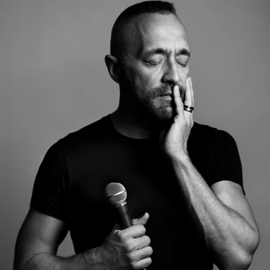 Black and white image of Theo Blackman who is holding a microphone, closing his eyes with his hand placed on his face