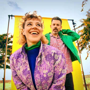 Two members of Tune-Yards outdoors in front of a bright yellow backdrop, surrounded by trees