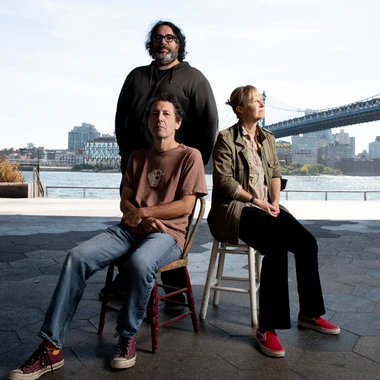 Three members of Yo La Tengo seated and standing on a waterfront plaza with a bridge in the background