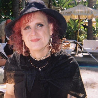 Portrait of Devra Hoff in a wide brimmed black hat, black lace top, and layered necklaces, smiling at a poolside setting with palm trees.