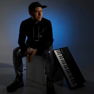 Portrait of Jason Lindner seated against a dark blue backdrop with a keyboard leaning beside him.