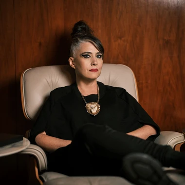 Kathleen Hanna wearing all black sitting in a beige leather Eames chair against a dark brown wooden wall.