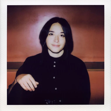 Portrait of Macie Stewart in a black button up shirt, smiling against a warm reddish backdrop.