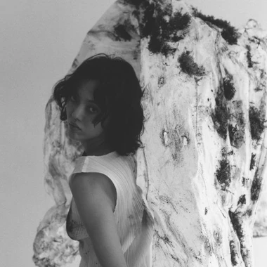 Black and white portrait of Rila Fukushima looking over her shoulder beside a large textured, moss patched sculptural form.