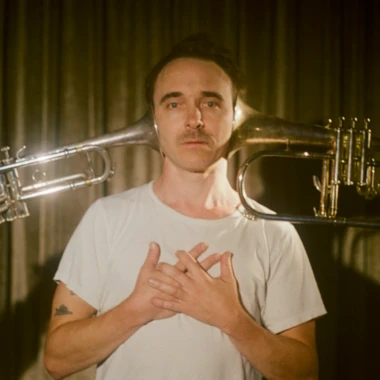 Portrait of Toth in a white t shirt, two trumpets flanking his head like wings, hands crossed over his chest.