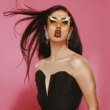 Bohenne Arreaux, drag queen, dressed in a black tube top, yellow tinted sunglasses, and their hair blowing in the wind against a pink backdrop.