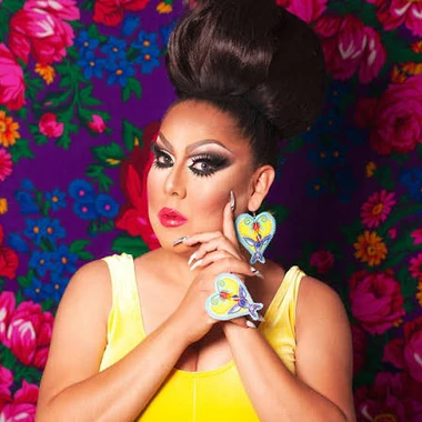 Lady Shug, drag queen, wearing yellow against a bright floral patterned fabric background.
