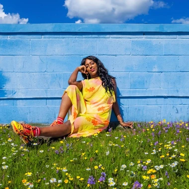 Passionfruit (Tana Yonas) the artist sitting against a bright blue wall sitting on grass wearing a yellow dress and smiling.