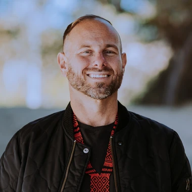 El G smiling wearing a red shirt with a black jacket against a blurred background