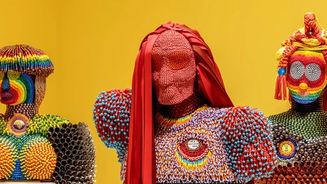 Image of three of Jeffrey Gibson's beaded and fringed multicolored human busts against a yellow background