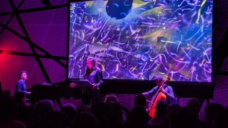 Three musicians performing on stage in front of a screen with artwork on it