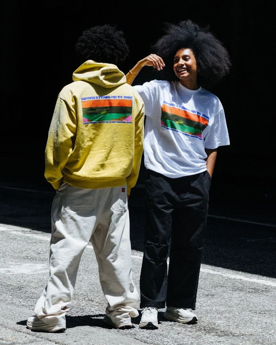  Two people stand outdoors in sunlight, one in a yellow graphic hoodie and cream wide-leg pants, the other smiling in a white graphic tee and black trousers.