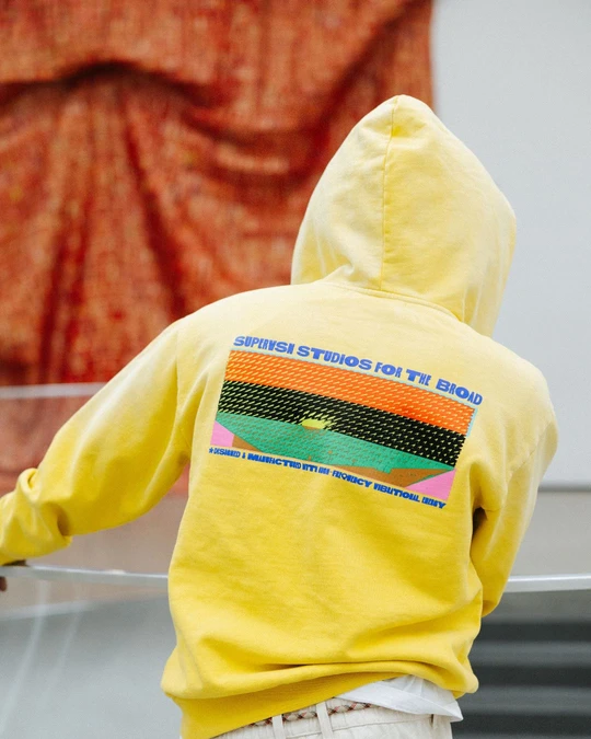 A person stands with their back to the camera in a gallery, wearing a yellow graphic hoodie, with a large red textile artwork visible on the wall behind them.
