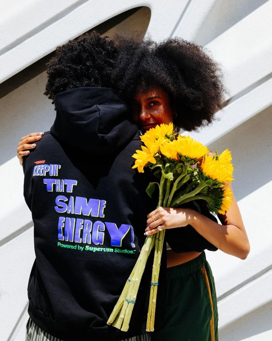 Two people embrace outside The Broad, one in a black graphic hoodie, the other smiling toward the camera while holding a bouquet of sunflowers.
