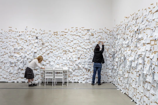Two visitors participating in Yoko Ono's artwork "my mommy is beautiful" by writing notes about their mother and pasting them on the walls