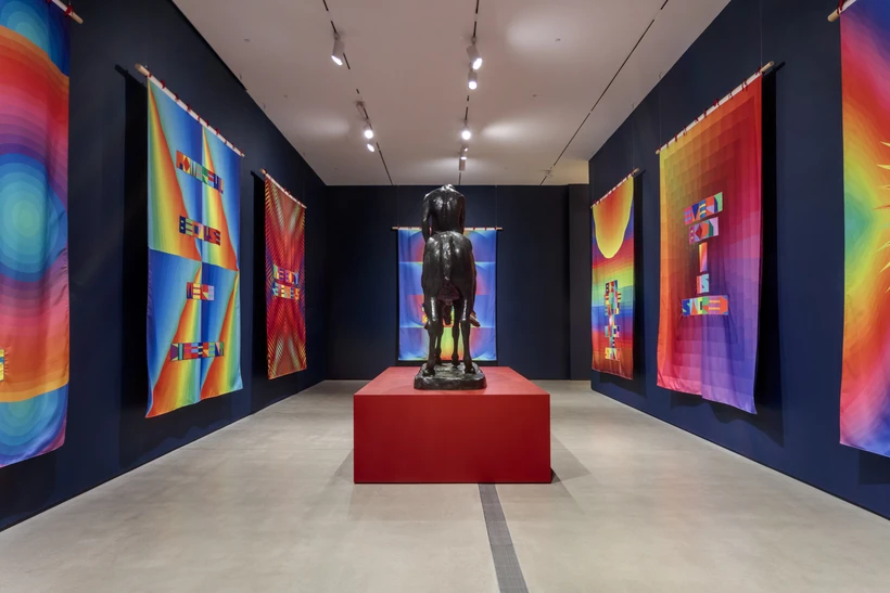 Bronze sculpture of a Native American man on a horse wearing moccasins in a dark blue walled gallery with 7 visible multicolored flags