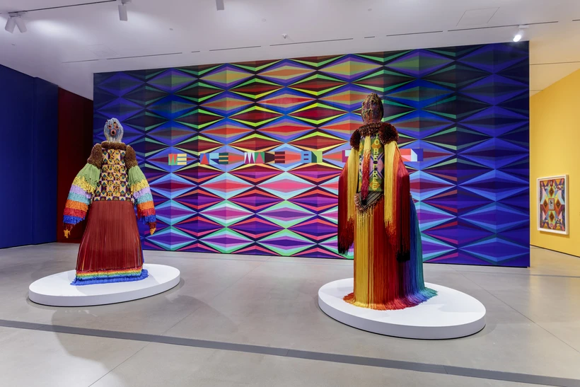 Multicolored beaded and fringed ancestral figure sculptures against a multicolored mural