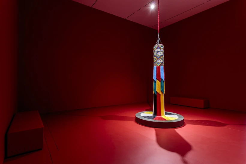 Multicolored, beaded, and fringed punching bag sculpture in an all red gallery