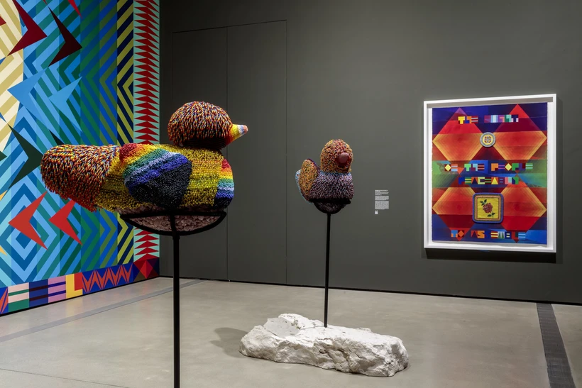 Two multicolored beaded bird sculptures from the side with a multicolored painting behind them to the right