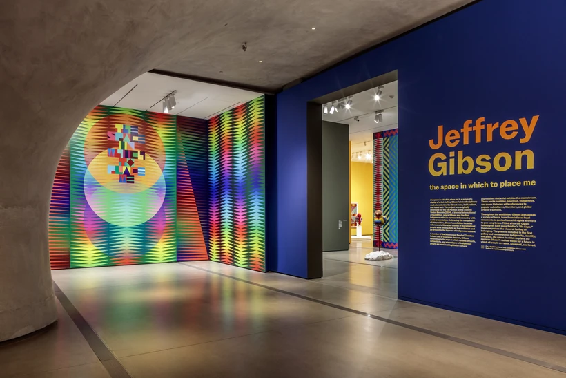 Blue Jeffrey Gibson title wall with orange and yellow text and multicolored mural inside The Broad