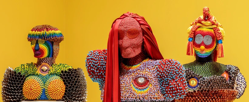 Image of three of Jeffrey Gibson's beaded and fringed multicolored human busts against a yellow background