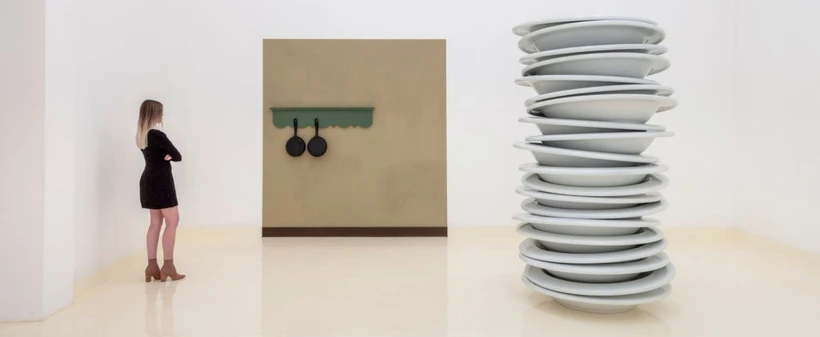 A woman in a black dress looking at Therrien's stack of plates artwork and an artwork featuring two black pans hung on a green panel against a brown wall