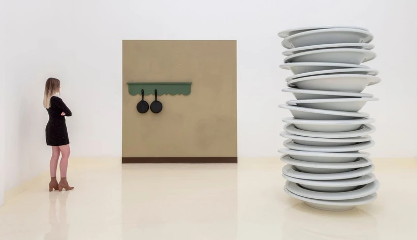 A woman in a dress standing in a gallery with a stack of oversized plates and a wall with two pots on it