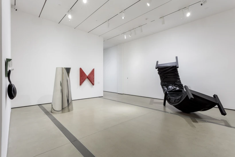 Installation image inside Robert Therrien: This is a Story, featuring an oversized silver pitcher and a curved black bed sculpture