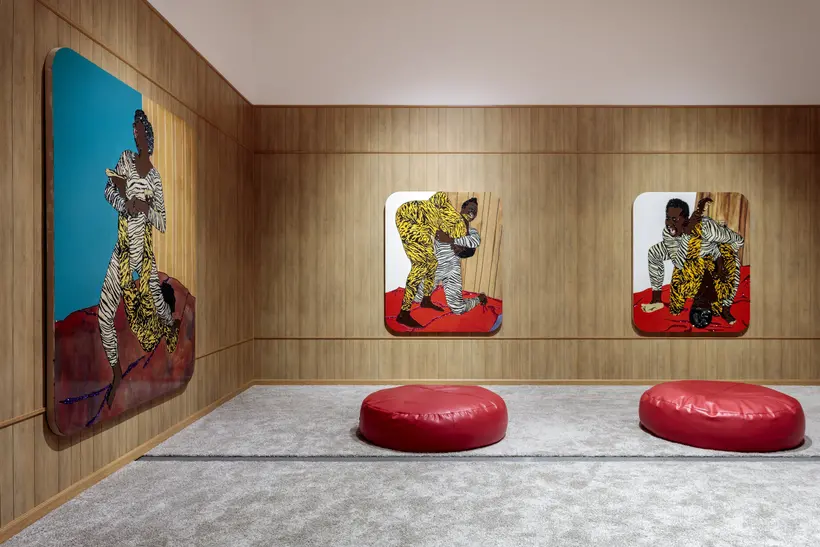 Gallery view of The Broad's Mickalene Thomas All About Love Exhibition featuring three paintings of women wrestling. The room has wood paneling with carpet and there are two beanbags.