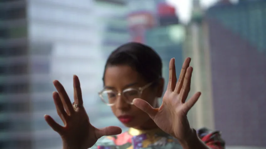 A woman wearing glasses holds her hands out in front of her. There are tall buildings in the background