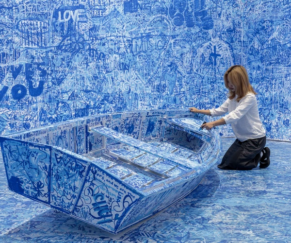 a visitor participating in Yoko Ono's artwork Add Colour (Refugee Boat) by drawing a sculpture of a boat with blue marker.