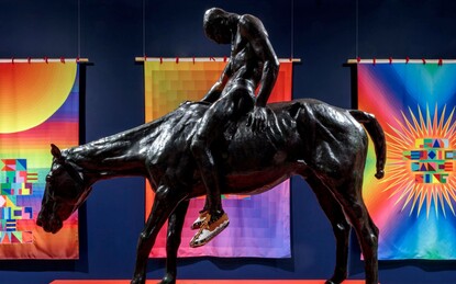 Bronze sculpture of a Native American man on a horse wearing moccasins in a dark blue walled gallery with 7 visible multicolored flags